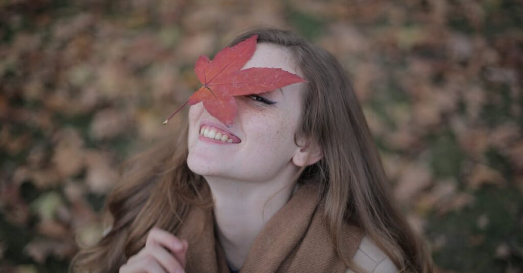 Smiling woman with maple leaf on face embracing fall season outdoors.