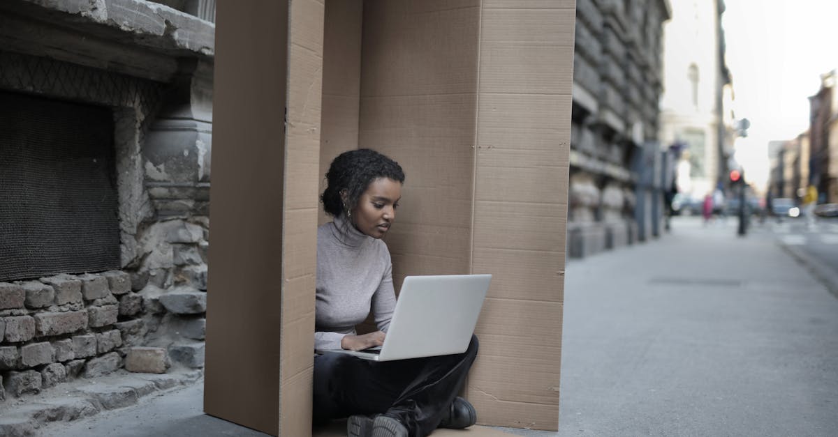 Young female introvert in casual clothes typing on netbook while sitting with crossed legs inside cardboard box located on asphalt road in city in afternoon