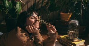 A woman with short hair smiling and enjoying dried flowers indoors surrounded by plants.