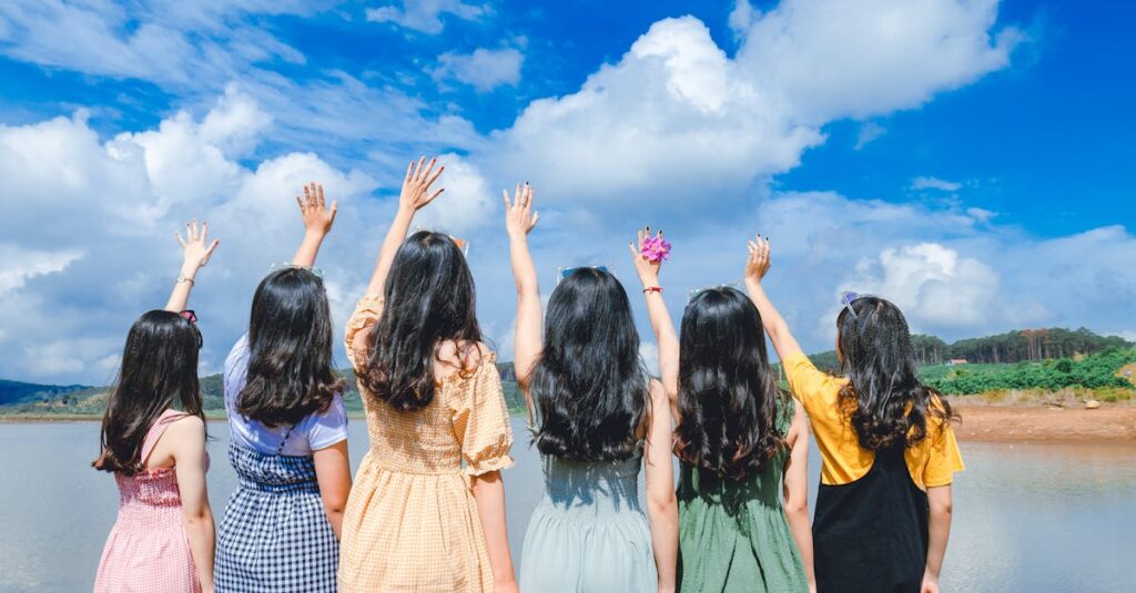 Group of young women with long hair raising hands by a lake under a clear blue sky.